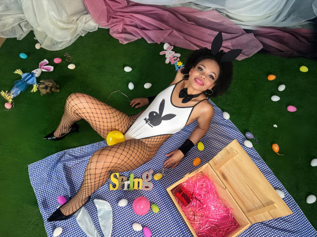 ChantalHaneke, with black bunny ears, fishnet stockings, and a white Playboy bodysuit, lies on a blue checkered blanket, legs spread, revealing her pussy, surrounded by Easter eggs and pink netting.