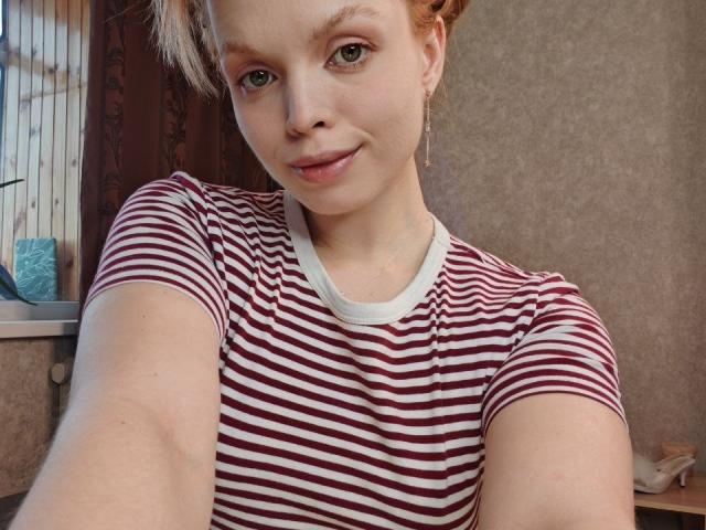 Sinteas with light skin, green eyes, and blonde hair, wearing a red and white striped shirt, smiling at the camera, holding the phone out. Her tits are small.