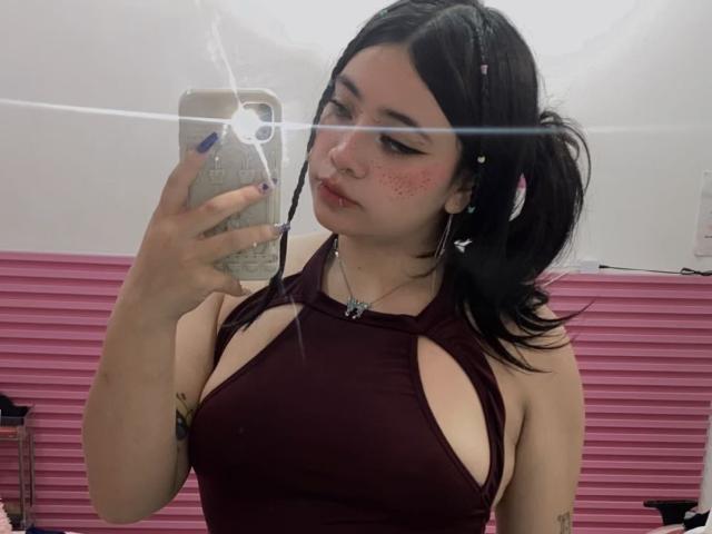 AmaraKazumi takes a mirror selfie with a smartphone. She has light skin, dark hair in pigtails, and wears a maroon halter top with large cutouts. She has freckles on her cheeks and a small nose ring. Her nails are painted blue. The reflection shows a bright flash, and the phone case is textured. She has a tattoo on her left forearm. The image is casual and focuses on her upper body and face.