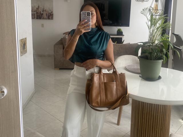 GalaMalkova with brown skin, wearing a teal blouse, white pants, and a brown leather bag, holding a phone, in a modern room with a green plant.