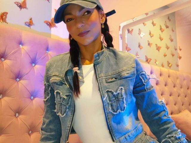 HannaPretty sits in a pink tufted booth. She wears a blue denim jacket with frayed edges and butterfly patches, a white shirt, and a blue cap. Her dark braids hang over her shoulders. Orange butterfly decals cover the background. Soft pink and purple lighting highlights her face and jacket. Her confident expression and slight smile are prominent. The image has a casual, modern style with a focus on her trendy, edgy look.