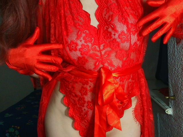 Aromalove wears a red lace robe with floral patterns, tied with a shiny red ribbon. Her pale skin contrasts with the bright red. She has long, red-gloved fingers, one on each side, touching the robe. The lace is semi-transparent, revealing the shape of her torso. The image focuses on her midsection and hands, with a textured wall and a bed with a blue patterned cover in the background. The robe is detailed and the gloves are shiny.