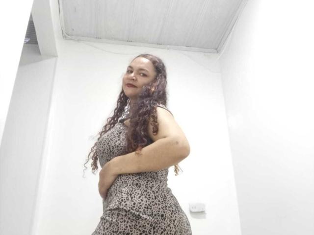 VivianBig has long curly brown hair, wearing a leopard print dress, standing with one hand on her hip, showing her ass. She has a slight, confident smile.