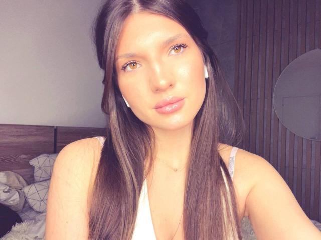 VivaDelavi is a young woman with long, straight brown hair and subtle highlights. She has light brown eyes, full lips with pinkish gloss, and a slightly flushed, shiny complexion. She wears white wireless earbuds and a white sleeveless top. The image is a close-up selfie, emphasizing her face and upper torso. The lighting is soft, highlighting her skin's smooth texture. Her expression is neutral, almost serene. The background is blurred, with a wooden headboard and a circular mirror. The composition is centered and focused on her face.