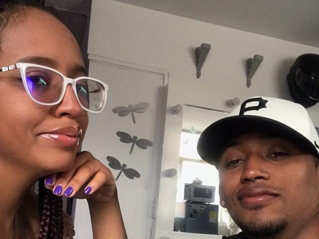 LilithAndAsmodeo with shiny lips, purple nails, and white glasses, next to a dude with a black and white hat, smiling. Her face is on the left, his on the right.