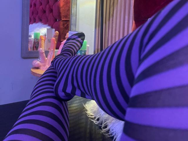 Close-up of GraceRedMiller's legs in purple and black striped socks, crossed and relaxed on a black surface. Reflection in a mirror shows a pink, tufted headboard and a cluttered shelf with sex toys and lotion. Soft purple and blue lighting creates a sensual atmosphere. Striped socks emphasize the texture and pattern. Slight shadowing adds depth. The image focuses on the legs and their striped pattern.