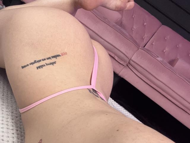 AlessiaNoire's pale, freckled ass is showing in pink thong, with a tattoo on her right butt cheek. Her back is slightly arched, and her feet are visible.