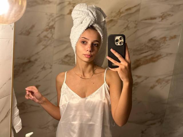 AishaBee with a white towel on her head, wearing a sheer white camisole, holding a black iPhone, standing in a marble-tiled bathroom, looking into the mirror.