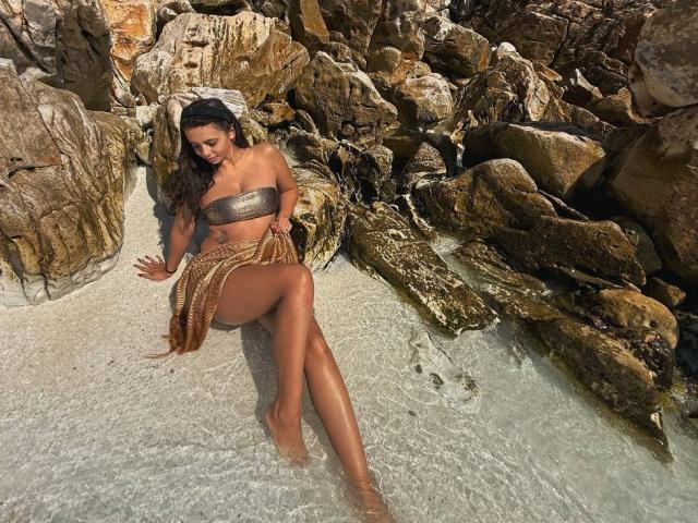 AishaBee with tan skin, long dark hair, small metallic tube top, brown fringe skirt, sitting on rocky beach, legs in clear water, showing her tits and ass.