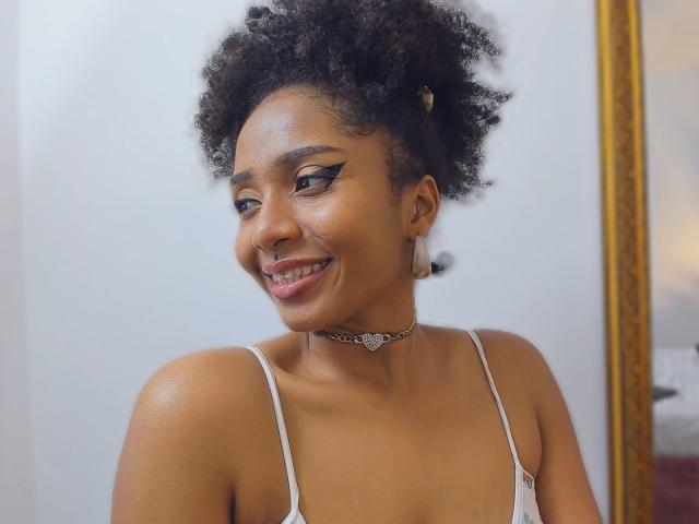 MeganWilliams has a curly, natural afro with a small gold hairpin. She's smiling slightly to the right, showing a bit of her teeth. She has dark brown skin and is wearing a white spaghetti-strap top. Her makeup includes black winged eyeliner. She's wearing a silver choker with a heart-shaped pendant. Her earrings are small, rectangular, and silver. The image focuses on her upper body and face. She has a subtle, confident smile.