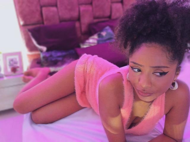 MeganWilliams is on a bed, lying on her stomach with her legs bent at the knees. She has dark curly hair in a high ponytail and wears a pink, fuzzy, sleeveless dress with a deep V-neck. Her makeup includes thick black eyeliner with a sharp wing. She has a nose ring and large white earrings. The bed has purple pillows and a pink, cushioned headboard. The image has a pink and purple hue, and the lighting is soft. MeganWilliams looks to the side with a slightly pouty expression.