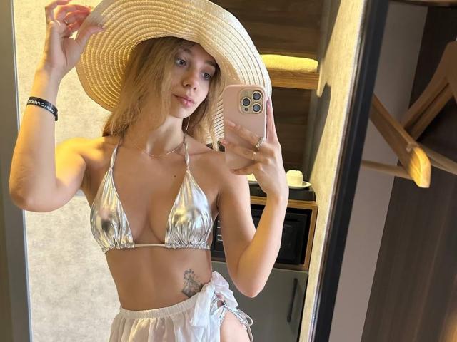 AishaBee in a silver bikini top showing small tits, wearing a straw hat, holding a pink phone, with a white skirt and a tattoo on her right hip.