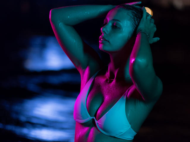 AishaBee with wet skin, arms raised, wearing a white bra, highlighting her big tits, in colorful blue and pink lighting, with a focused, sexy expression, standing by water.