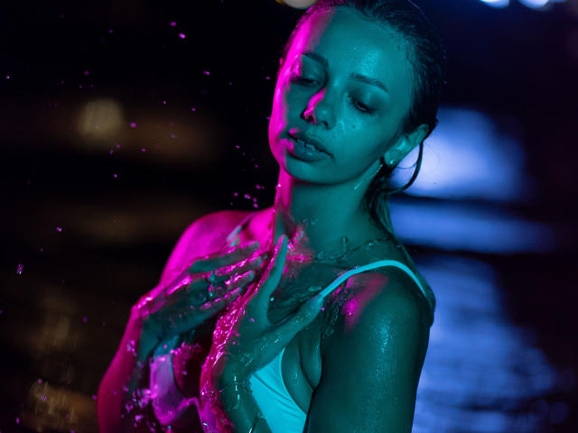AishaBee with wet blonde hair, blue-green light, wearing a white tank top, water droplets on her chest, eyes closed, lips slightly parted, neon pink and blue light.