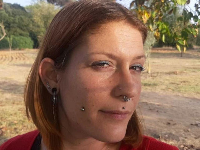 NinfaDeseo-hot has light brown skin, red shoulder-length hair, multiple facial piercings (nose and lower lip), and is wearing a red shirt. She's outdoors with a grassy background.