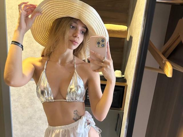 AishaBee with small tits in a shiny silver bikini top, holding a straw hat, wearing a white skirt, and a phone with three cameras, showing a tattoo on her hip.