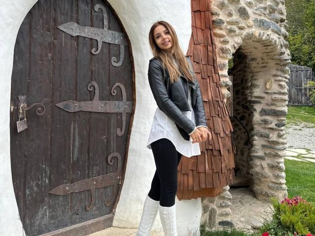 AishaBee stands in front of a wooden door with iron hinges, wearing a black leather jacket, white shirt, black pants, and white knee-high boots. She has long brown hair.