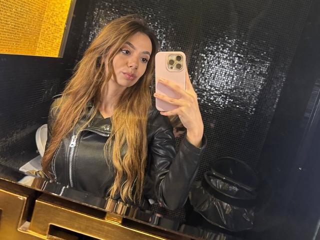 AishaBee with long brown hair, black leather jacket, and pink phone, taking a mirror selfie, showing her tits, in a shiny black-tiled bathroom.
