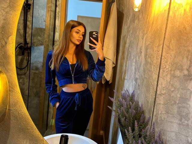 AishaBee with long brown hair, blue velvet crop top and pants, showing a tattoo on her right side, taking a mirror selfie in a dimly lit bathroom.