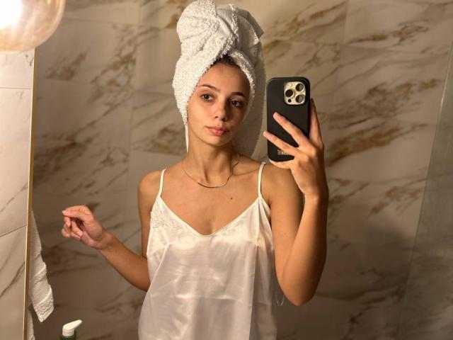 AishaBee with a white towel on her head, wearing a sheer white camisole, taking a mirror selfie with her phone, showing her tits and a slim, attractive build.