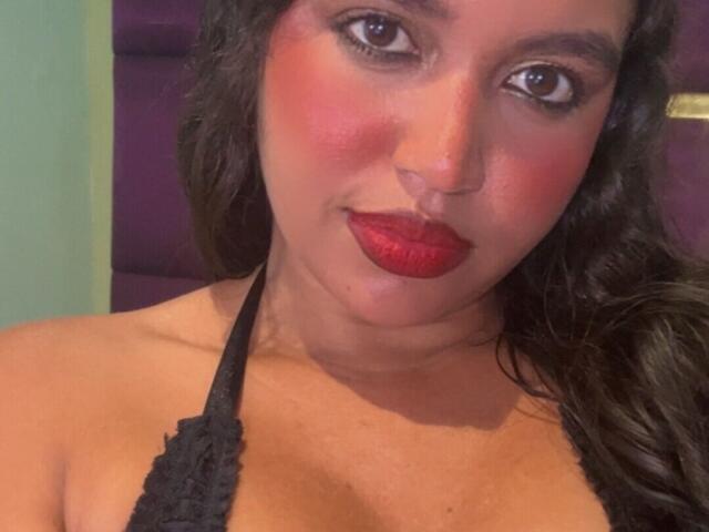 JeimyCampos with red lipstick, dark wavy hair, black lace halter, flushed cheeks, and a slight smile. Her brown eyes are looking directly at the camera. Boobs visible.
