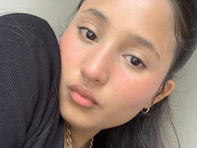 Ximenitax's face, with puffy pink lips, brown eyes, and light skin, wearing a black ribbed shirt, has a gold chain necklace and small hoop earrings, looking slightly to the side.