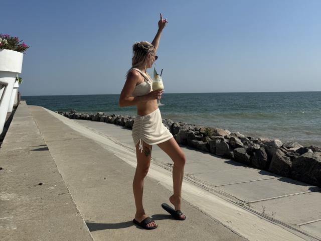 A photograph of XinitaBoom standing on a concrete walkway by the sea. She's wearing a beige bikini top, a white skirt, and black flip-flops. She holds a green drink with a straw in her right hand and points upward with her left hand. She has long blonde hair, sunglasses, and a small gold bracelet on her right wrist. The sea is in the background, with a rock breakwater and clear blue sky. XinitaBoom is in profile, facing right.