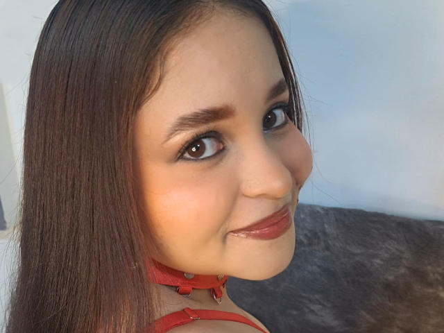 SamyMancini with glossy brown eyes, straight brown hair, red lipstick, and a red choker, smiling at the camera. Her ass is not visible. Her tits are not visible.