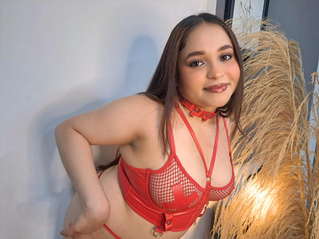 SamyMancini with red fishnet lingerie, showing her tits, and a red choker, smiling at the camera, standing next to pampas grass. She has brown hair and light skin.