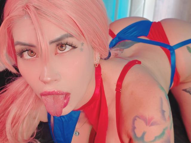 Close-up photo of Kkole with pink hair, brown eyes, and red lipstick. Kkole is on all fours, tongue out, wearing red and blue lingerie. Visible tattoos on Kkole's shoulder and upper arm. Kkole's right ear has a small hoop earring. Kkole's hair is slightly messy, covering part of their face. Bright, even lighting. The image focuses on Kkole's face, upper body, and tongue. Kkole's expression is playful. The lingerie straps are blue with red accents. Kkole's skin is fair. The tattoo on the shoulder is a colorful butterfly. The tattoo on the upper arm is partially visible.