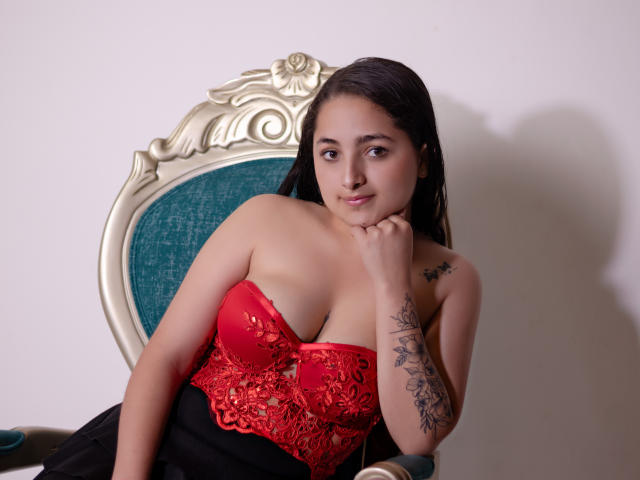 VickyGiacomo, a young woman with medium-length dark hair, sits on a vintage green and gold chair. She wears a red lace strapless top that accentuates her medium-sized breasts and a black skirt. Her right hand rests on her chin, and she has a subtle, confident smile. A floral tattoo is visible on her right forearm. The image has a watermark in the bottom left corner. Her skin tone is light olive, and her expression is relaxed. The lighting is soft, highlighting her facial features and the intricate lace pattern of her top.