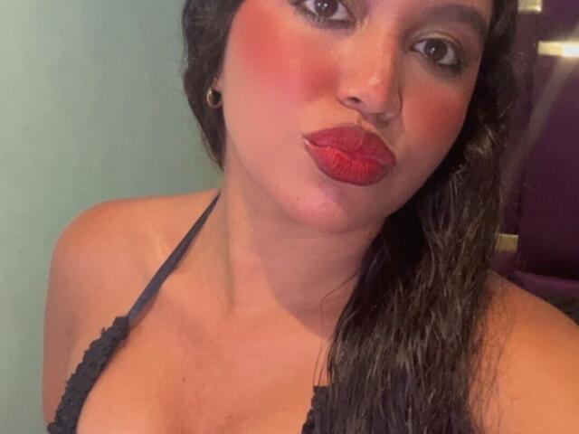 JeimyCampos with red lipstick, pouty lips, and black halter top, showing off her tits, with wet dark hair, and greenish background. Her ass is not visible.