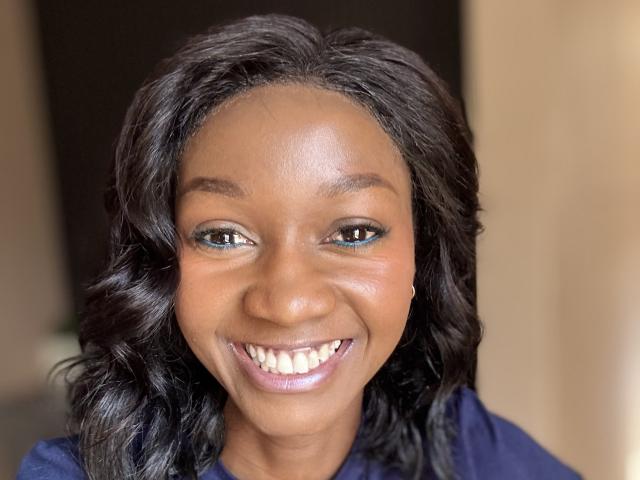 Mysteryslady's dark brown, wavy hair frames her smiling face with blue eyes, subtle eyeliner, and glossy pink lips. She's wearing a dark blue shirt, showing her white teeth.
