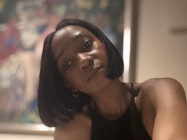Mysterylady with dark skin, short black bob, small hoop earrings, black tank top, slightly tilted head, and a serious expression, looking directly at the camera.