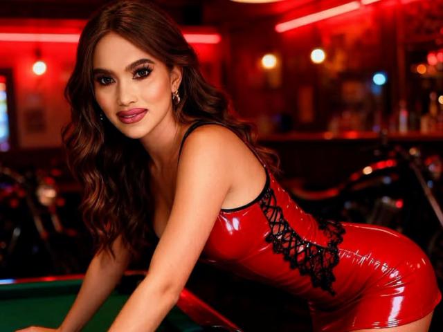 A sexy, brown-haired HornyCumTs with wavy hair leans over a green pool table, wearing a tight, red latex dress with black lace-up sides. She has dark, smoky eye makeup and pink lipstick. The background is blurred, with red lighting and a bar. Her right hand is on the table, and her left arm is out of frame. The image is sharp, with a focus on her face and dress. She looks directly at the camera with a slight, confident smile.