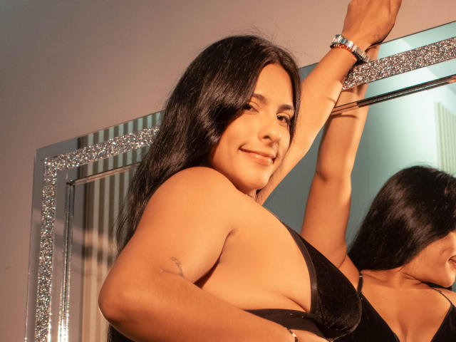 Nina Bloom, con cabello negro largo, sonriendo, levantando los brazos, vistiendo un sostén negro, reflejo en un espejo brillante, luz solar, piel bronceada, tatuaje pequeño en el brazo izquierdo, pulsera en la muñeca derecha..
