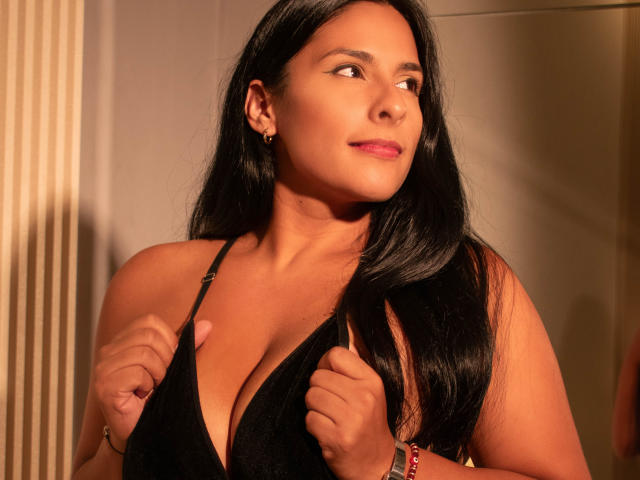Nina Bloom, con lunghi capelli neri, pelle abbronzata e rossetto rosa, tiene le spalline di un abito nero, mostrando il seno, guarda di lato con un leggero sorriso, indossando un braccialetto rosso..