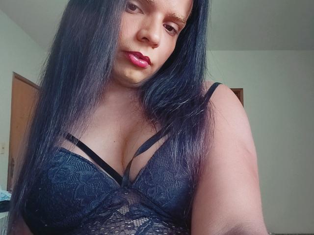 Aripietra with long black hair, dark lipstick, and a black lace bra, showing her tits, standing in a room, looking down at the camera.
