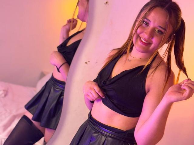 AnnikaBlake stands in a room, smiling at the camera. She has light brown hair in pigtails, wearing a black sleeveless top, and a black pleated skirt with a shiny texture. She lifts her top, exposing her midriff. Her reflection in a mirror shows her from the back. She wears black thigh-high stockings. Warm, pinkish lighting highlights her face and the mirror. Her expression is cheerful, and she holds a pigtail with her right hand.