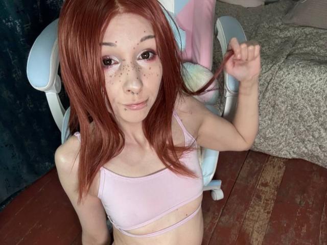 HannaCool with red hair, freckles, and piercings, wearing a pink sports bra, holding her hair, sitting in a chair, on a wooden floor, looking at the camera.