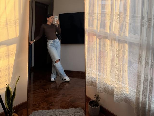ThaliaAres with small tits, wearing a brown long-sleeve top, ripped blue jeans, and white sneakers, taking a mirror selfie, standing in a sunlit room with lace curtains.