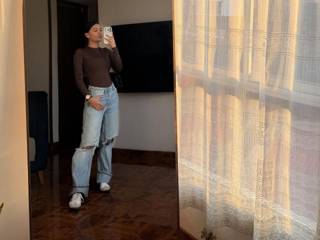 ThaliaAres has a slim, cute ass in ripped blue jeans, a brown sheer long-sleeve top, and white sneakers, taking a mirror selfie with sunlight through sheer curtains.
