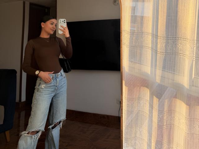 ThaliaAres with brown top, ripped blue jeans, standing, holding phone, mirror selfie, dark hair in bun, gold watch, black bag, sunlight through curtain, wooden floor, flat ass.