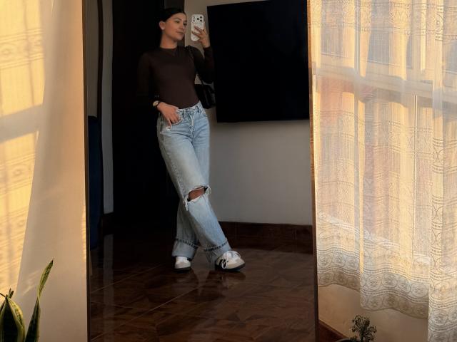 ThaliaAres, standing in a mirror, wearing a brown long-sleeve top, ripped blue jeans, and white Adidas sneakers, holding a phone, with sunlight and curtains in the background.