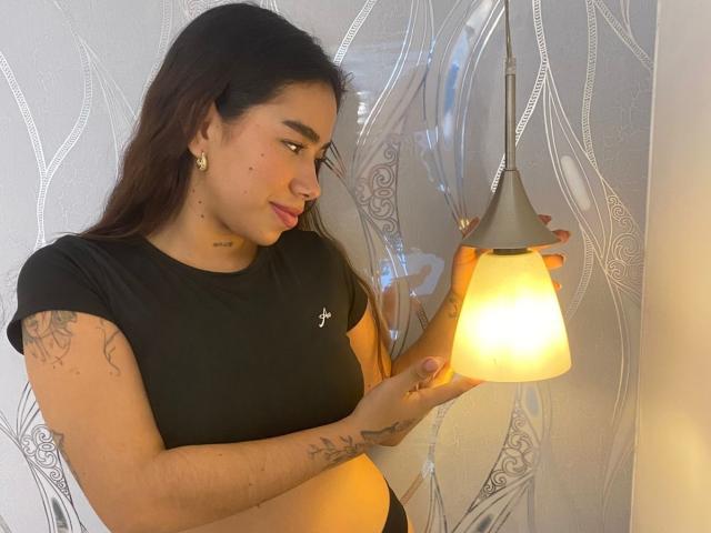 Saalome69, a young woman with long dark hair, stands in a room, holding a warm, yellow-hued pendant lamp. She wears a black, short-sleeved, crop top that reveals her midriff and small, gold earrings. Her right arm, adorned with a tattoo, is raised to adjust the lamp. She has a small tattoo on her lower right cheek and another on her right forearm. The wall behind her has a wavy, patterned design. The image has a soft, warm light.