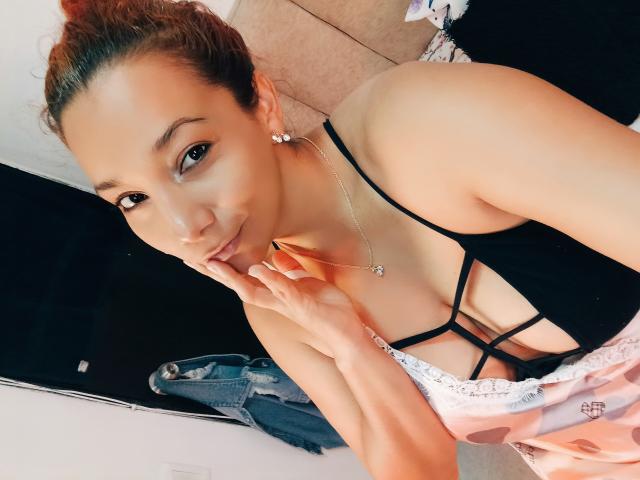 A close-up photograph of AmberDemons with light brown skin and curly auburn hair, wearing a black strappy top that reveals cleavage. She has small, rounded earrings and a delicate necklace. AmberDemons is looking at the camera with a slight smile, her right hand touching her chin. She's wearing a pink, lace-trimmed garment. The background includes a black mirror and a folded denim jacket on a beige floor. The image has a casual, candid style.