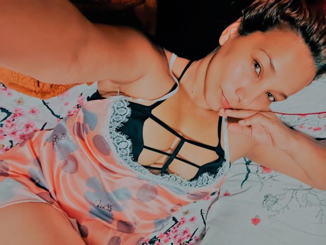 AmberDemons lies on a bed, taking a selfie. She has light brown skin and brown hair, with her head slightly turned to the camera. She wears a pink and gray polka-dotted nightgown with black lace trim and a black strappy bra. Her right hand touches her chin, and she has a subtle, confident smile. The bed has a floral-patterned sheet. The image uses warm, natural lighting, highlighting her facial features and the texture of her nightgown. The composition is close-up and slightly angled.