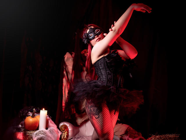 SanitaCooper with black mask, corset, and fishnet stockings, arms raised, standing in red light, surrounded by candles, pumpkin, and skull on dark surface. Her ass is prominent.