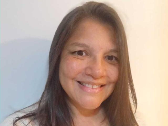 EstelaHot has brown eyes, straight brown hair, and a smile showing white teeth. She's wearing a white top with visible freckles on her cheeks and nose.