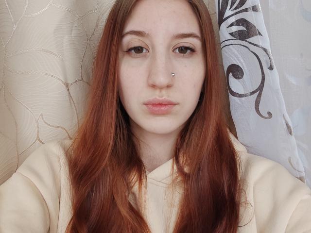 AuroreProvosty has pale skin, long red hair, a nose piercing, and is wearing a cream-colored shirt, looking straight at the camera with a neutral expression.